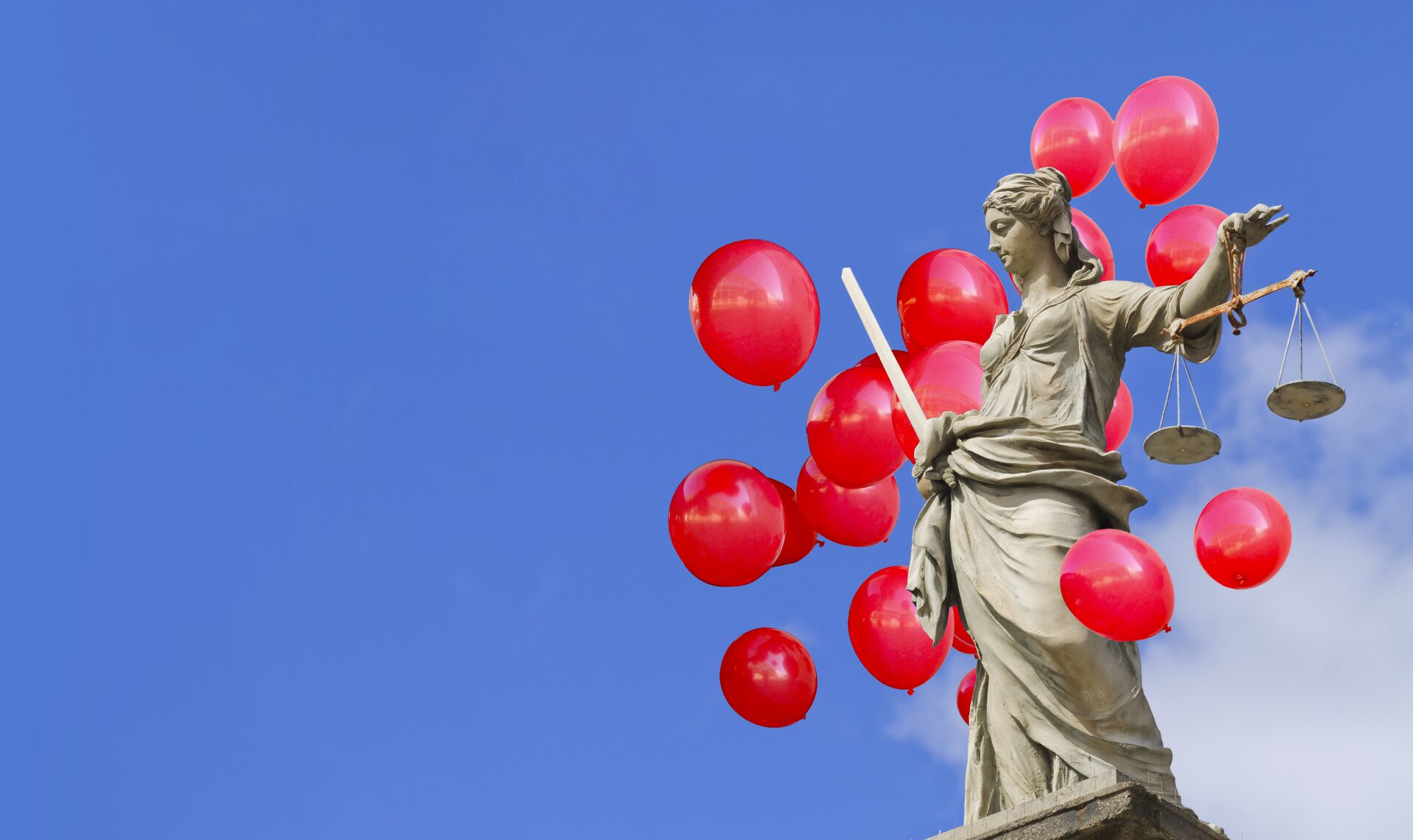 A statue of Lady Justice holding scales, surrounded by red balloons against a blue sky.